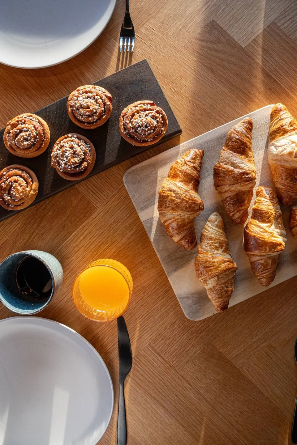Cinnamon buns, croissants, coffee and orange juice on an oak table in morning sun