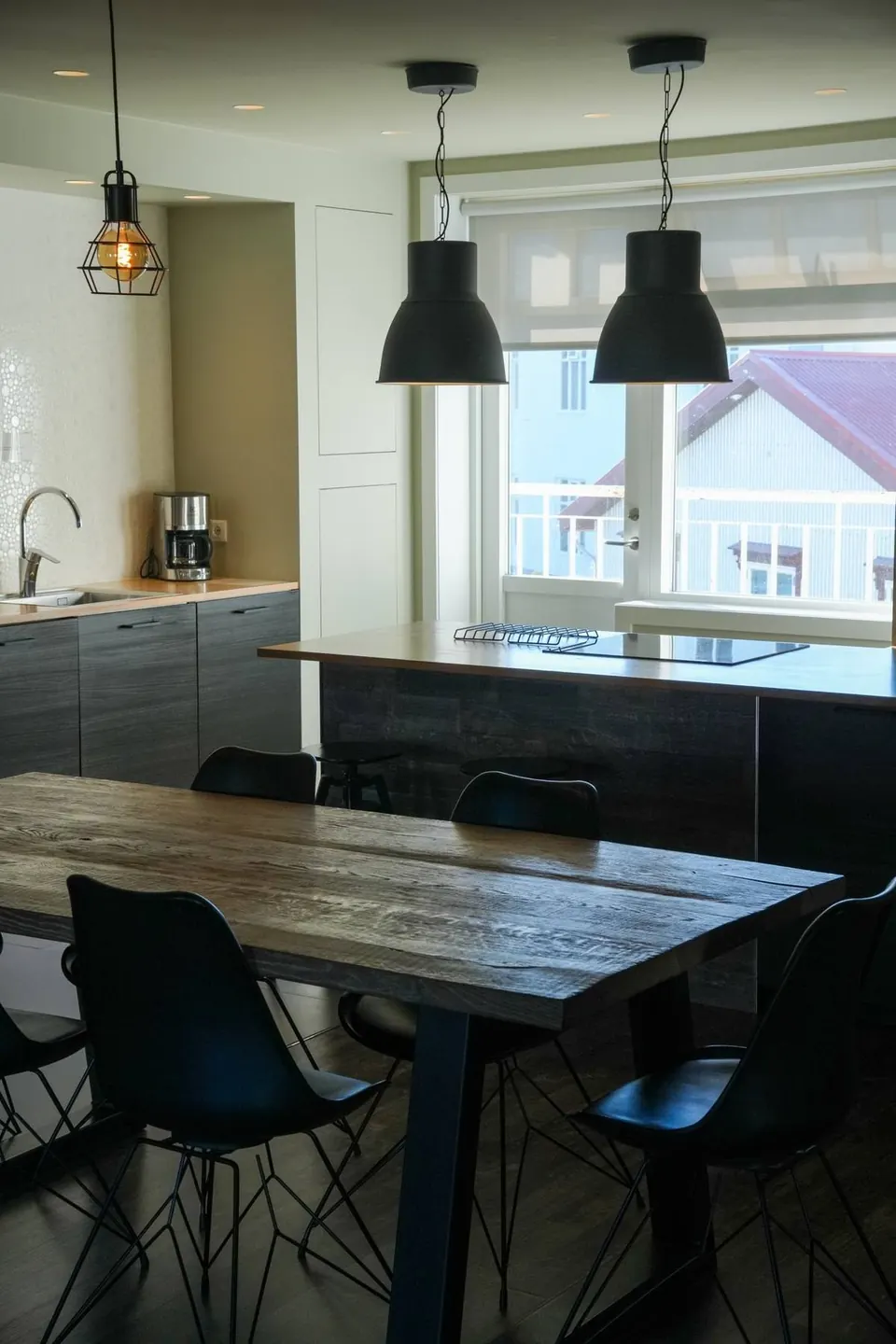 Kitchen and dining table under black pendant lamps, with a balcony door and town roofs outside